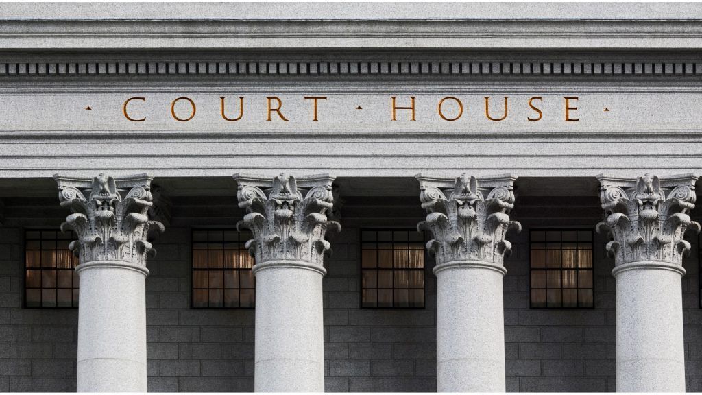 The stone facade of a courthouse with gold lettering and four ornate, classical columns in front of rectangular windows.