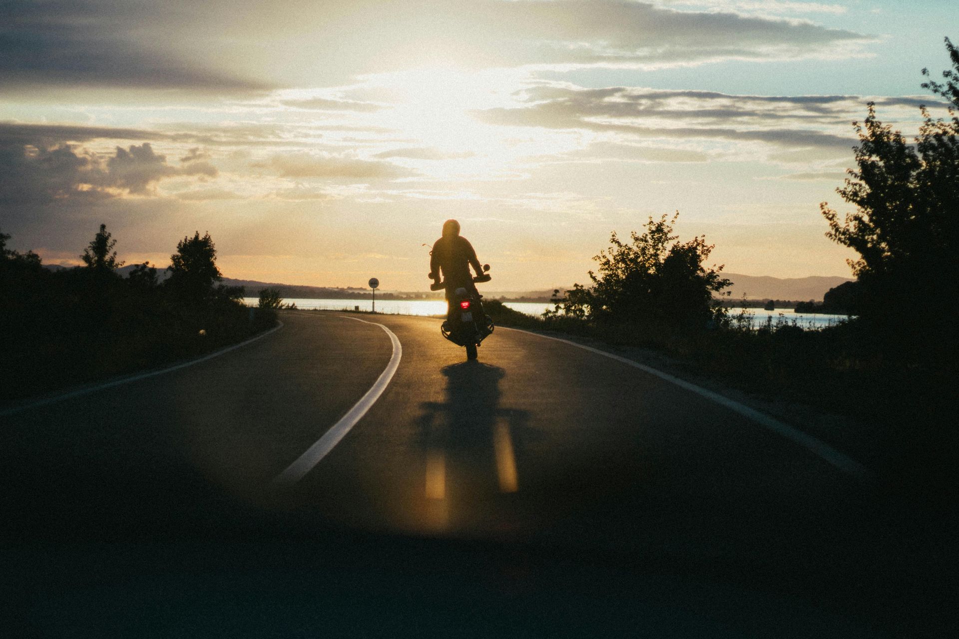 A lone motorcyclist riding down a paved road toward a sunset over a distant body of water.