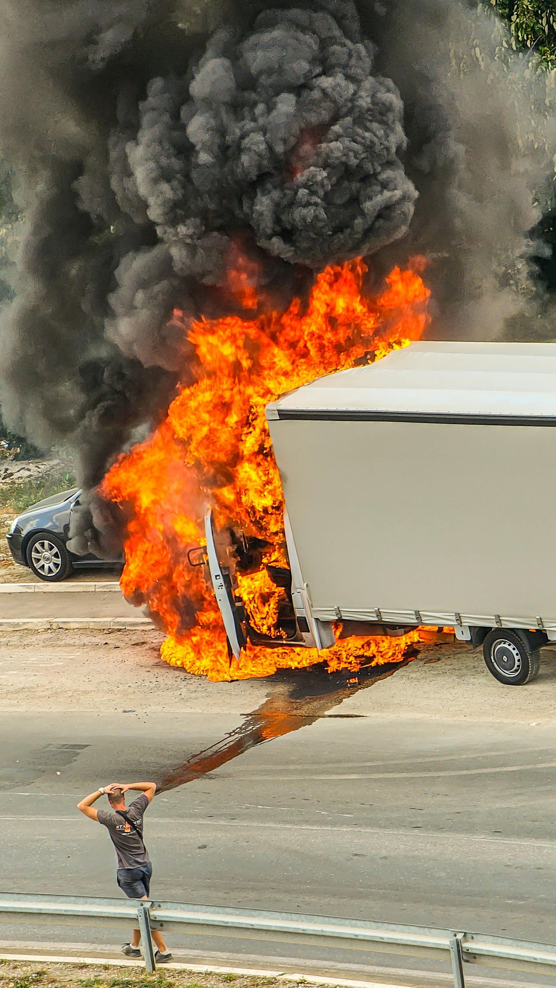 A trailer engulfed in bright orange flames and thick black smoke on a road, with a person standing nearby watching.
