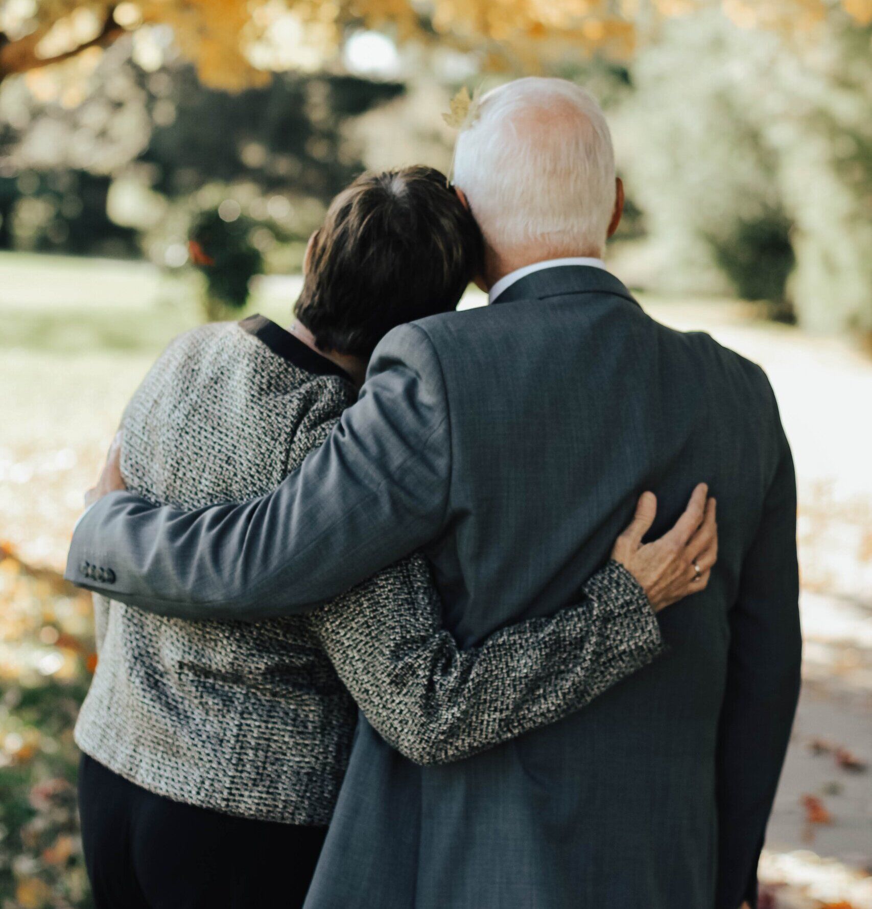 Two people standing side-by-side with arms around each other, looking at a blurred outdoor park setting.