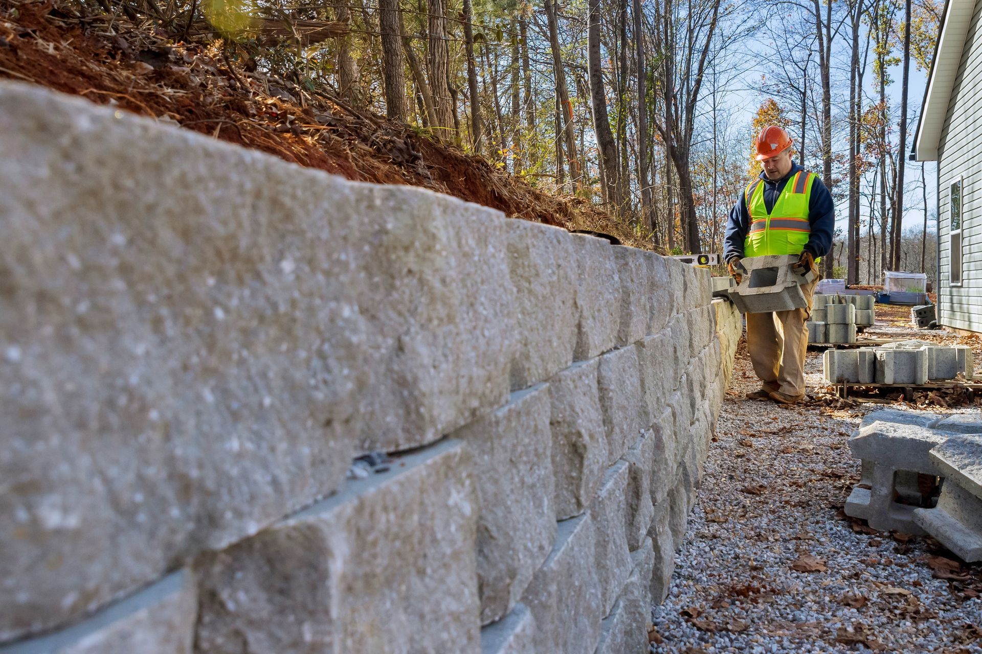 Stone retaining wall under construction beside a wooded path, with a worker in a safety vest.
