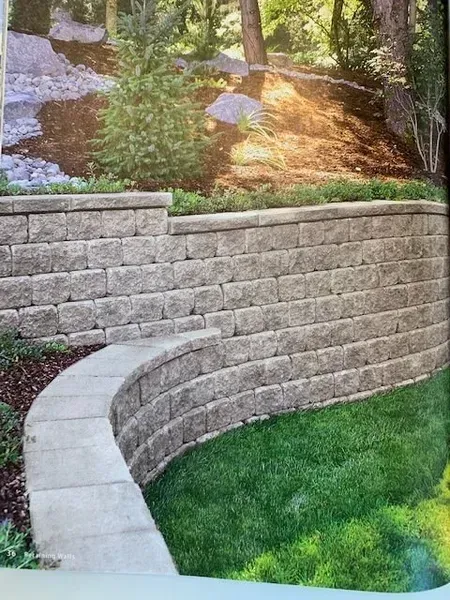Curved stone retaining wall beside a green lawn and wooded slope with rocks and a small stream