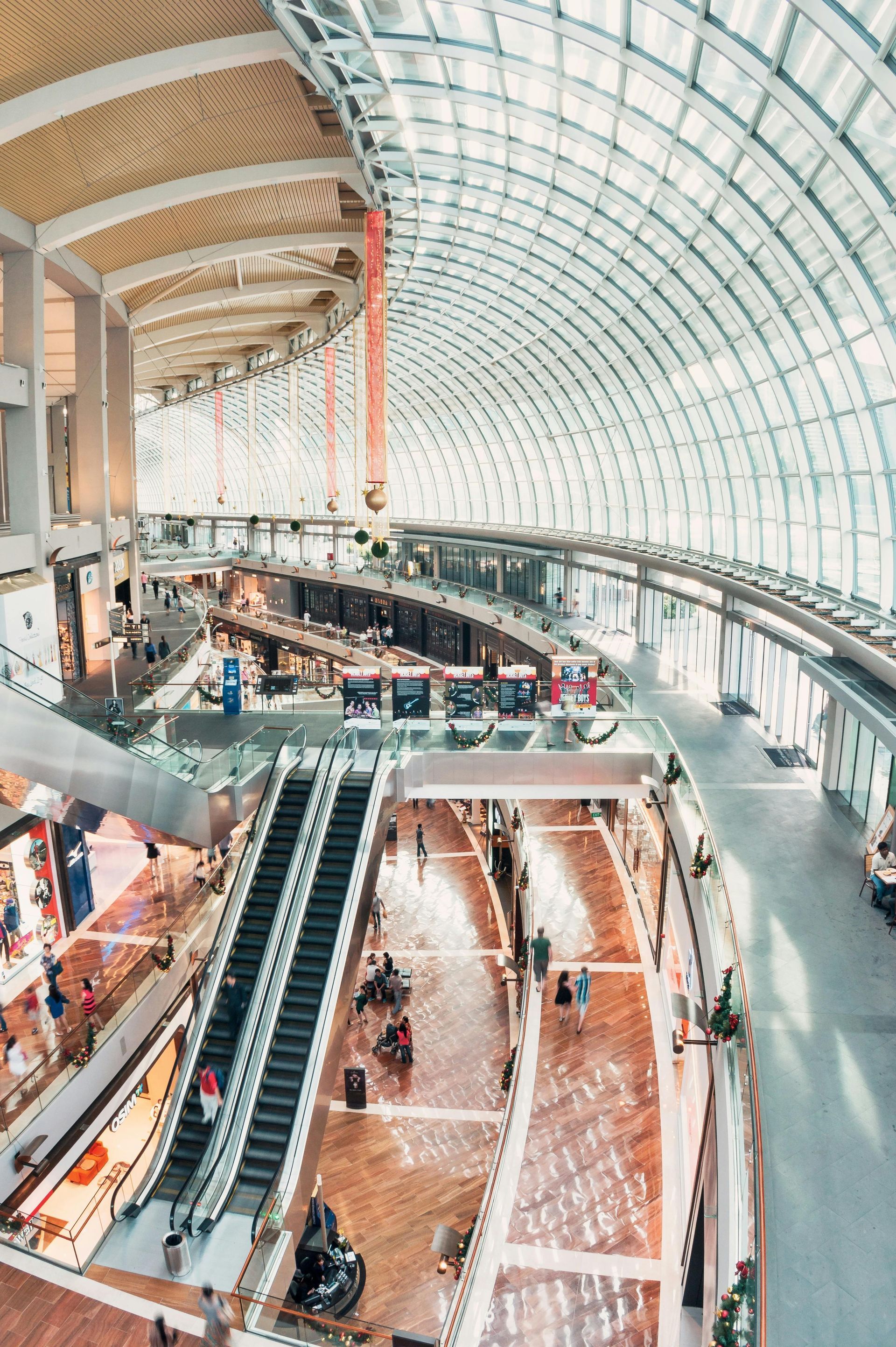 Multi-level shopping mall interior with curved glass ceiling and escalators.