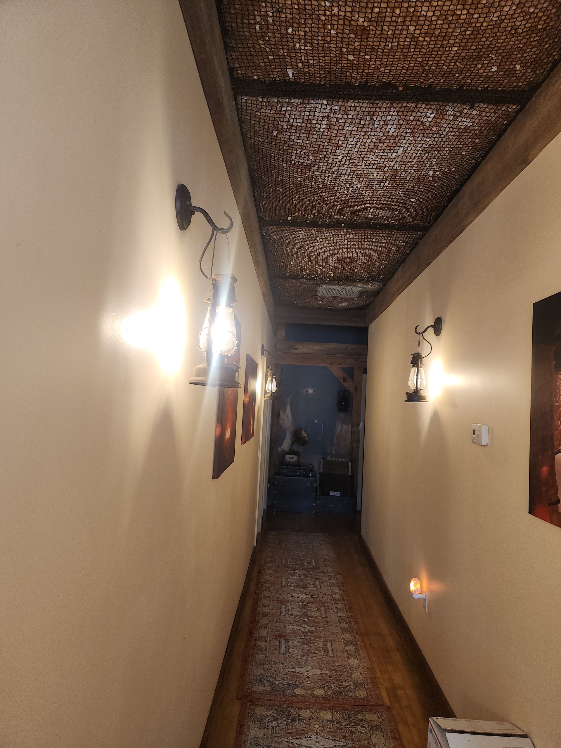 Narrow hallway with tan walls, wooden ceiling, ornate lighting, and a patterned rug.