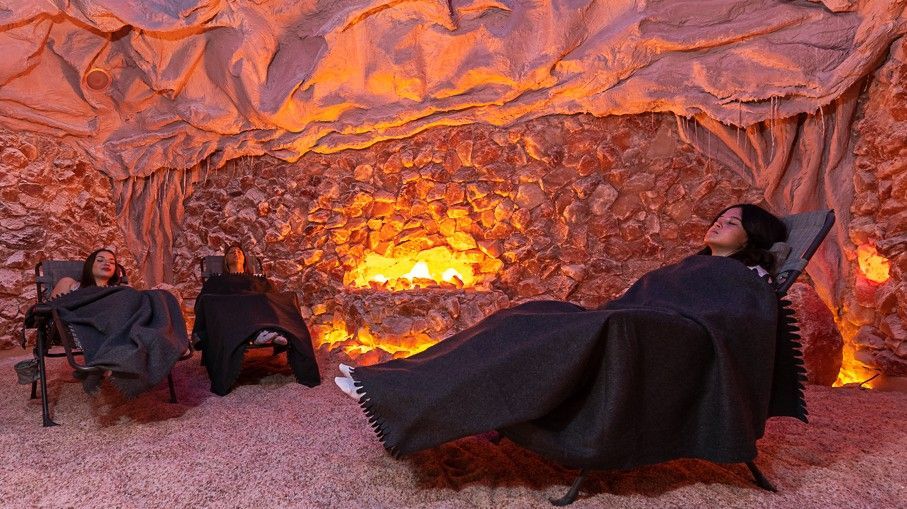Three people relax in a dimly lit salt cave. They recline on chairs, covered in blankets, amidst glowing orange salt formations.