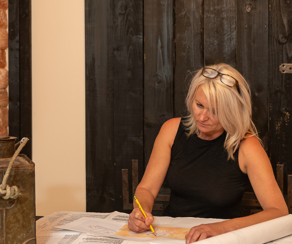 A person with blonde hair and glasses on their head sketching with a yellow pencil on paper at a table near a dark wall.