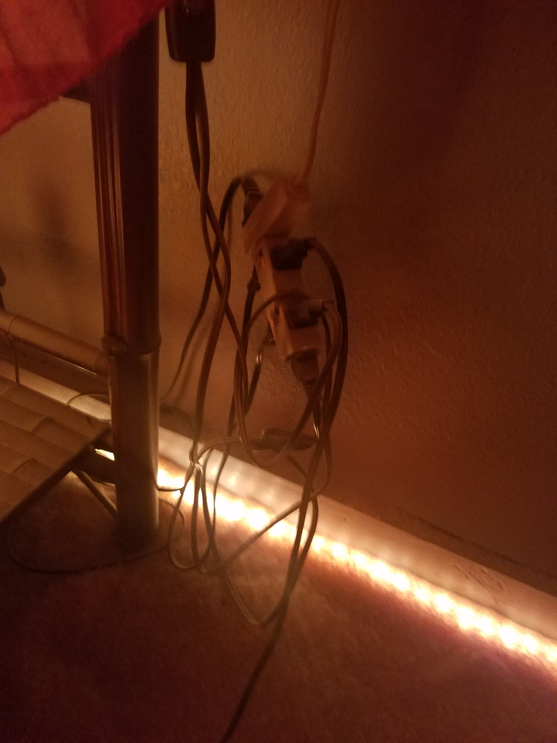 Tangled power cords plugged into wall outlets, near a wooden pole and an LED strip light along the floor.