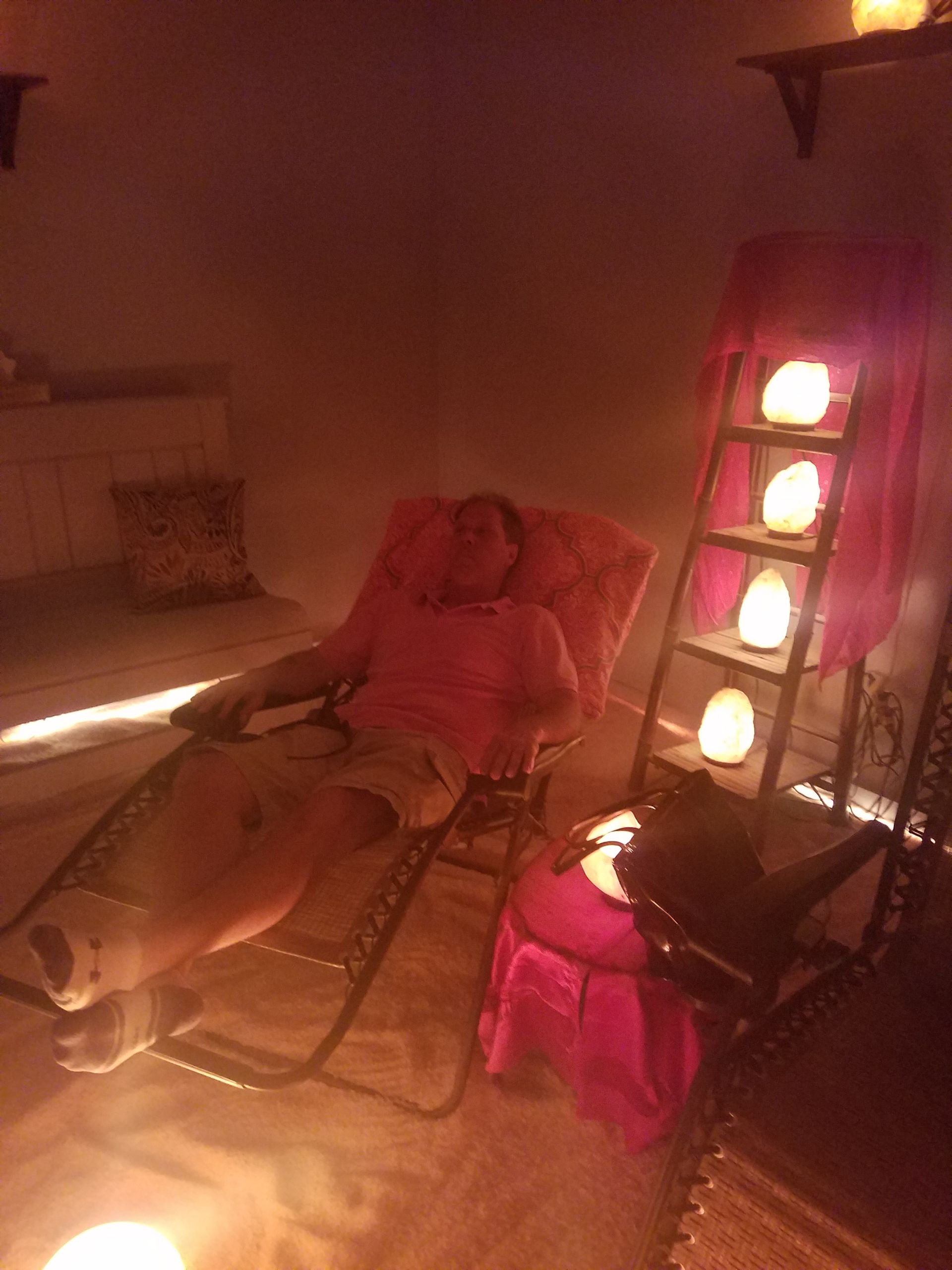 Man relaxing in chair inside a room with salt lamps, pink curtain, soft lighting.