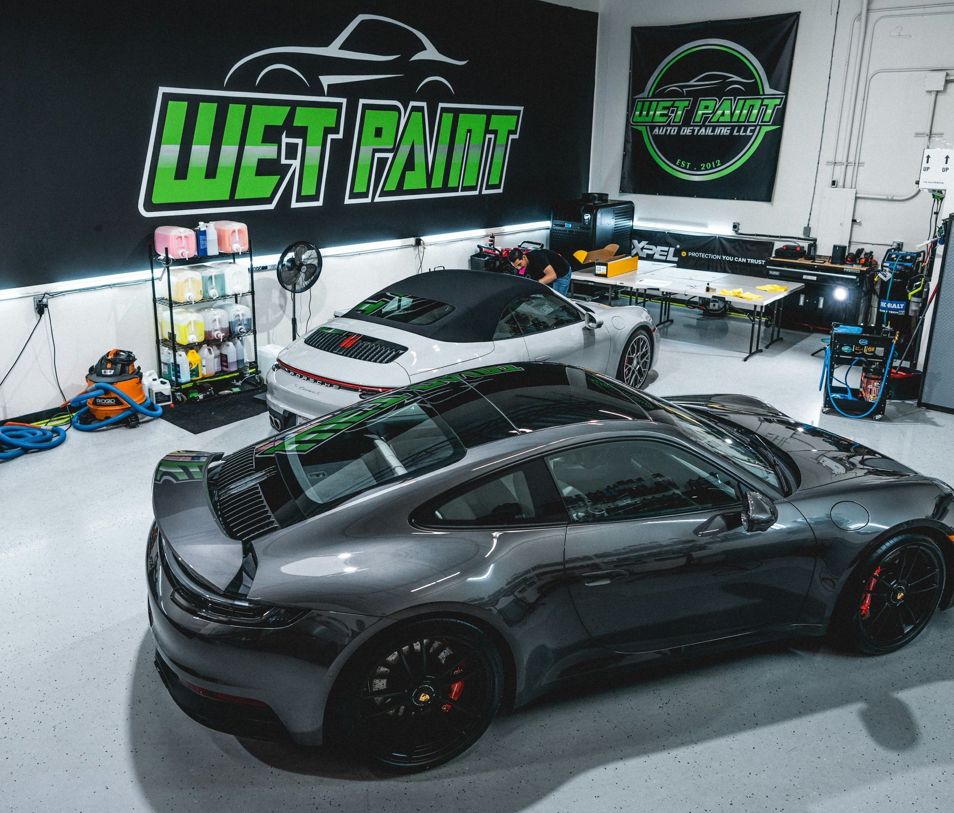 Two luxury sports cars inside a car detailing shop; shop features “Wet Paint” logo.