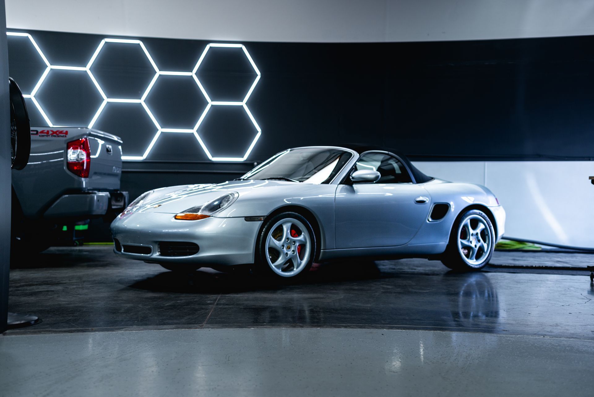 Silver Porsche Boxster convertible in a garage with hexagon-shaped lights on the wall.