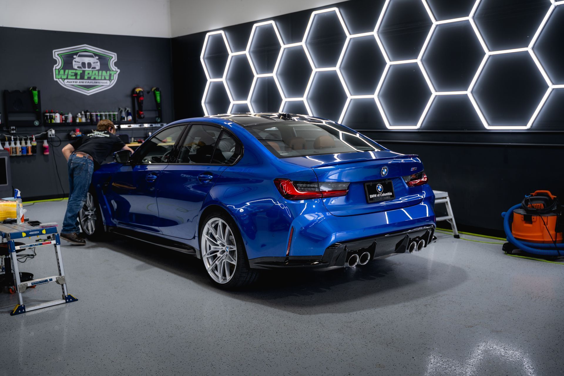 A blue BMW sedan inside a garage, two people working on it. Honeycomb lights on the wall.
