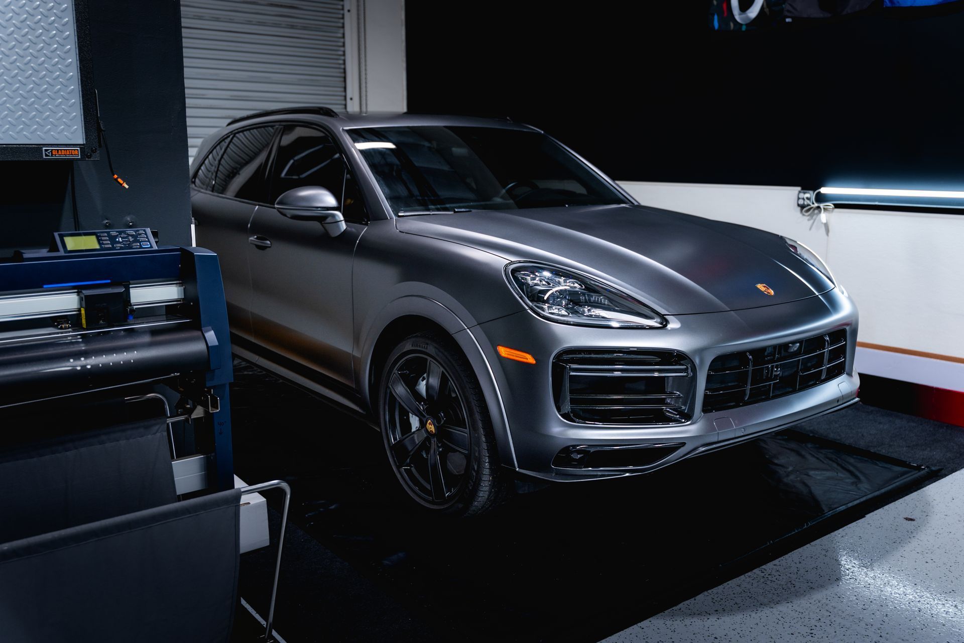 Gray Porsche SUV parked in a garage. Black wheels, headlights on.