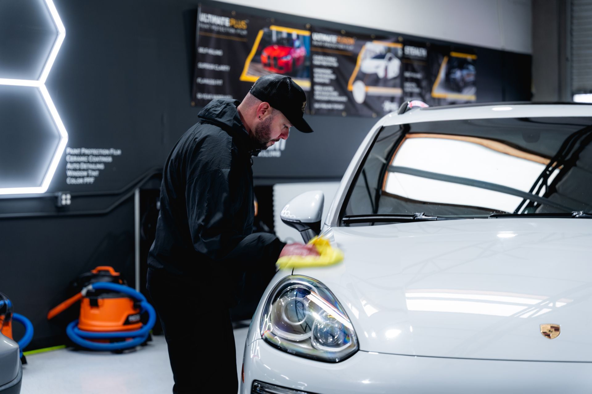 A person in a black jacket and cap polishes the hood of a white Porsche in a well-lit, professional automotive workshop.