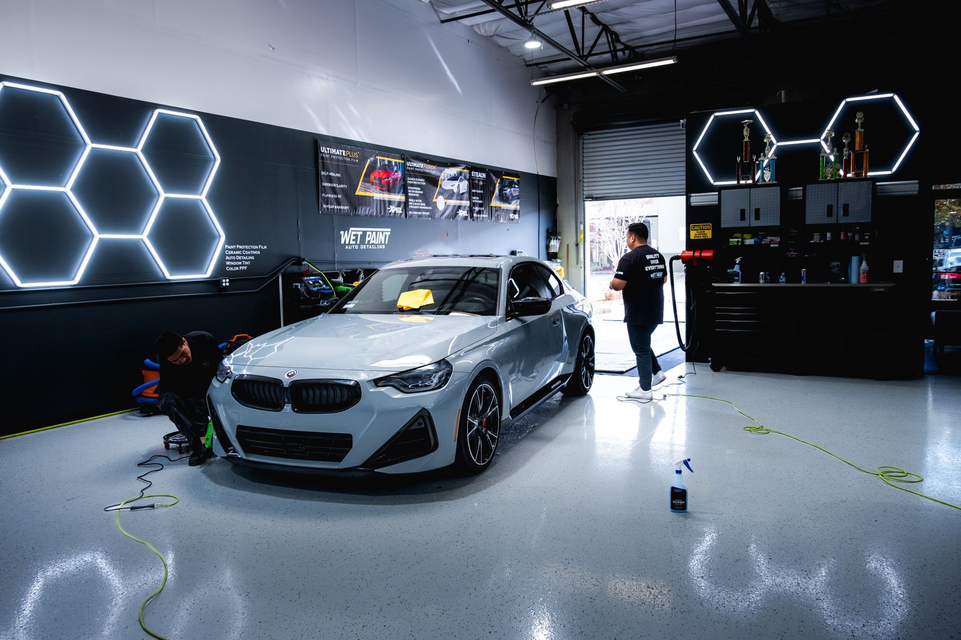 A gray BMW being worked on inside a garage. A person walks toward the car, which is in front of hexagon lights.