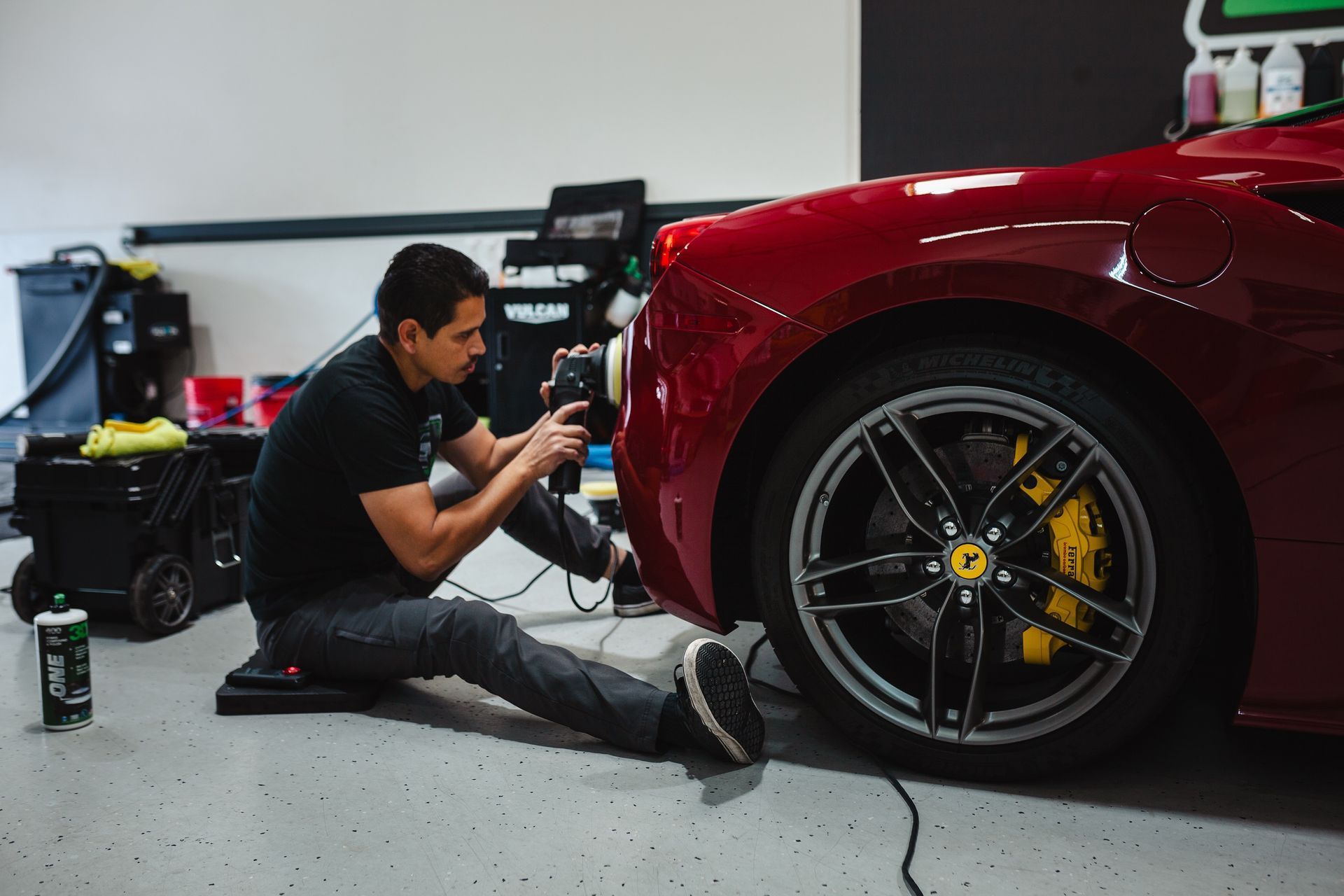 A man is kneeling down next to a red sports car in a garage.
