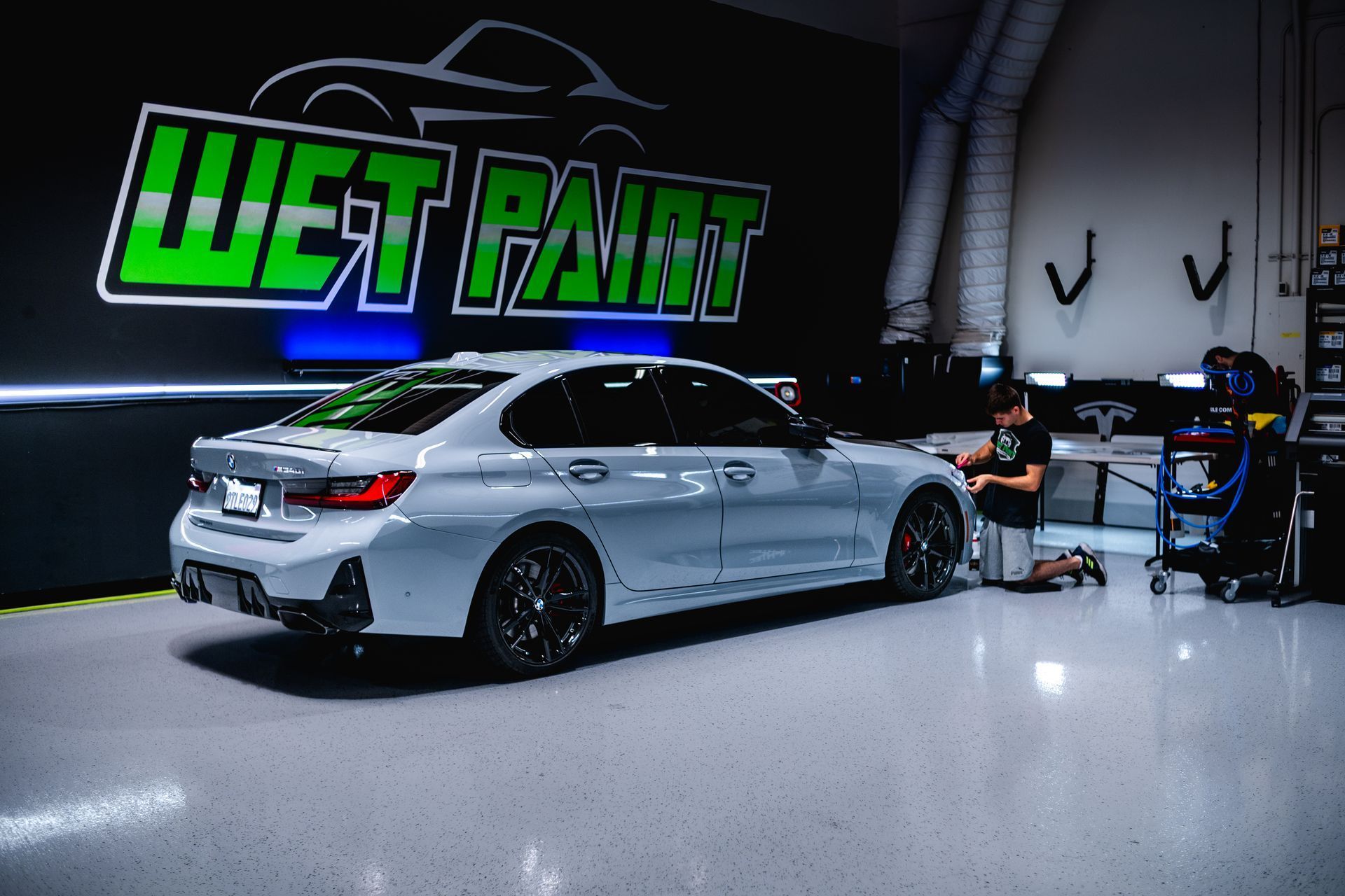 A gray BMW sedan in a car detailing shop. A person is working on the car. The shop sign reads 