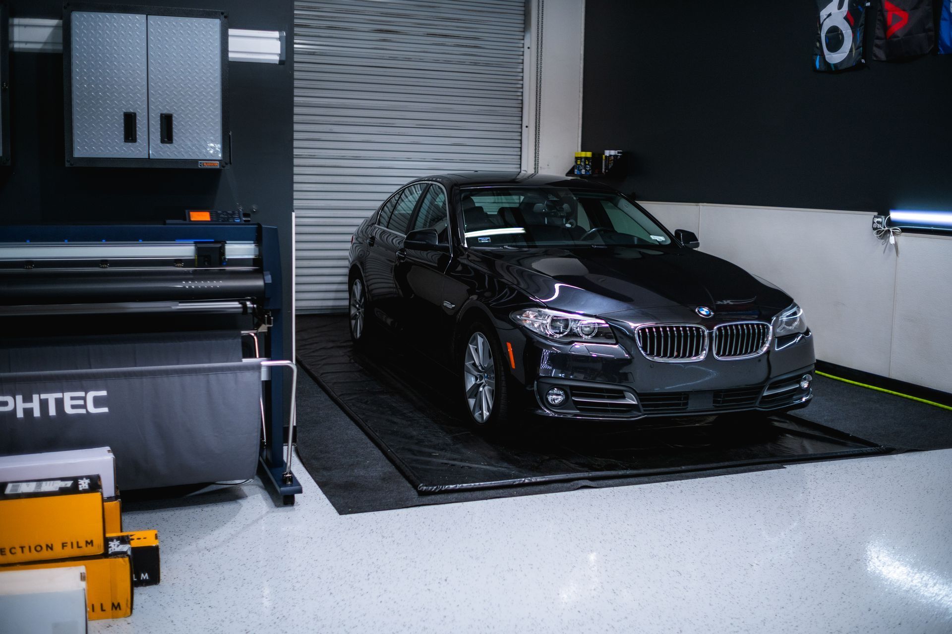 Black BMW sedan inside a shop. Vinyl wrap printer and cabinets visible, near a closed garage door.