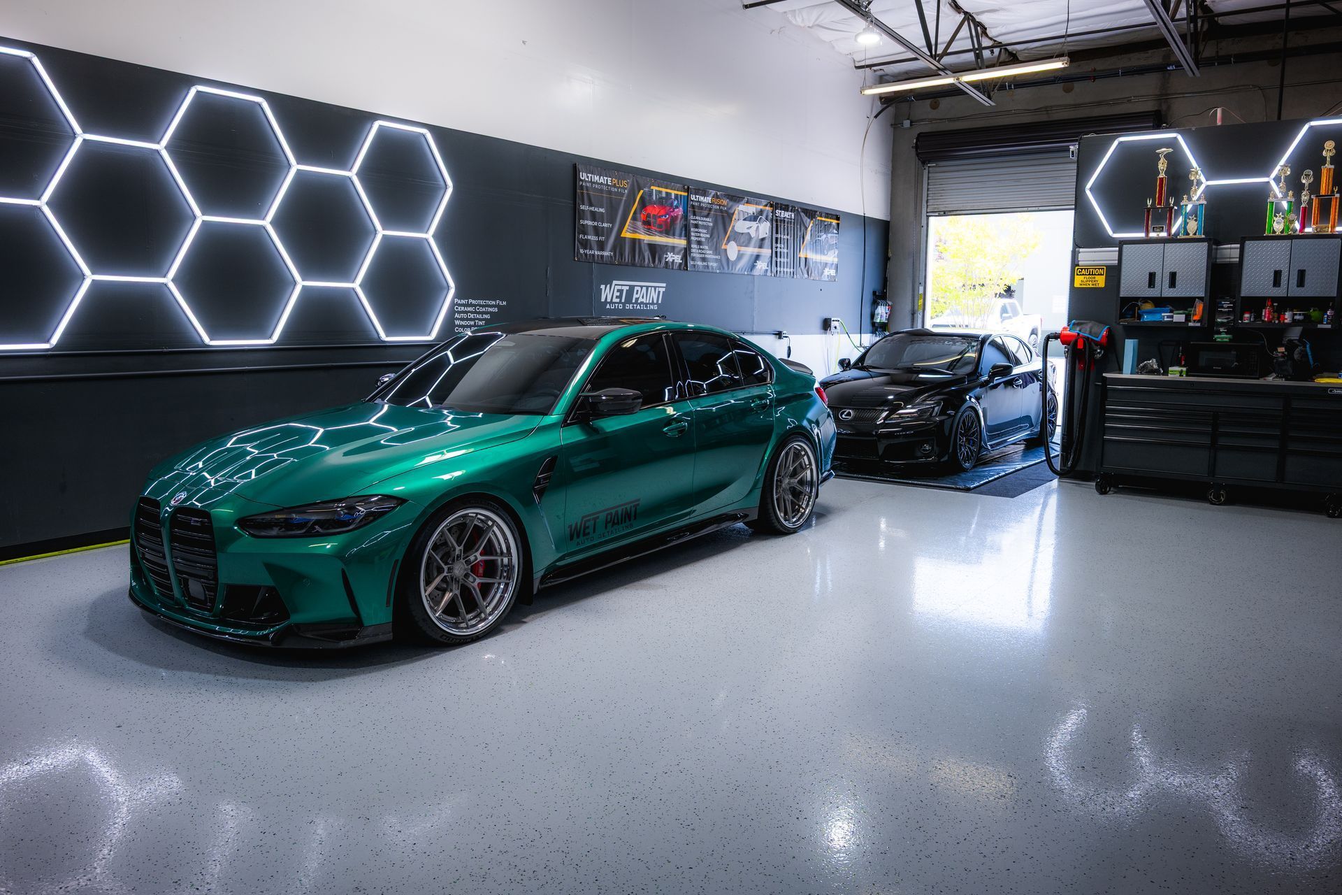 Emerald green car in a car detailing garage with a black car. Hexagon lights on the wall.