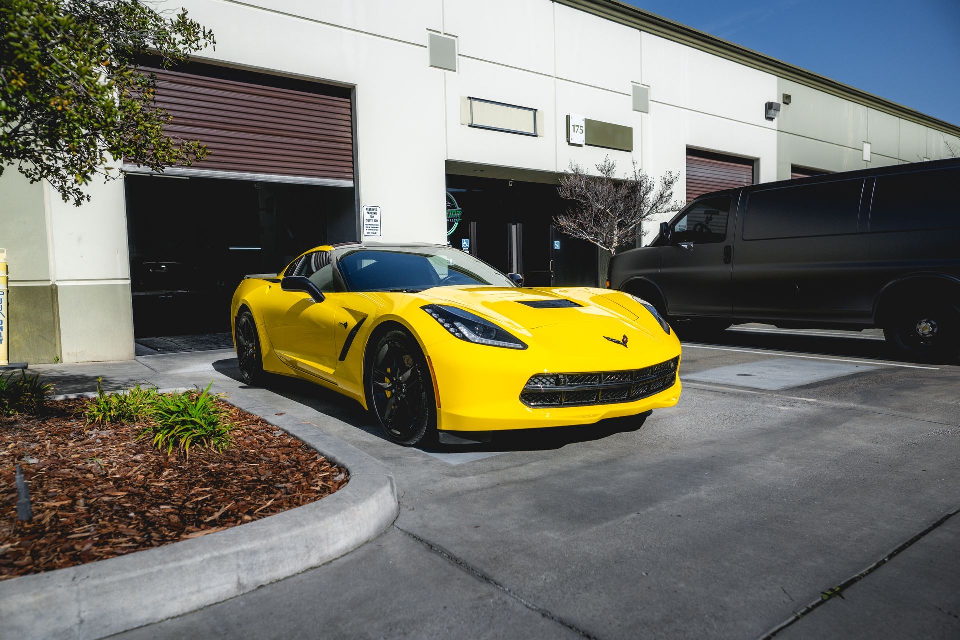 Yellow Corvette parked in front of a building with a partially open garage door. Black van on the right.