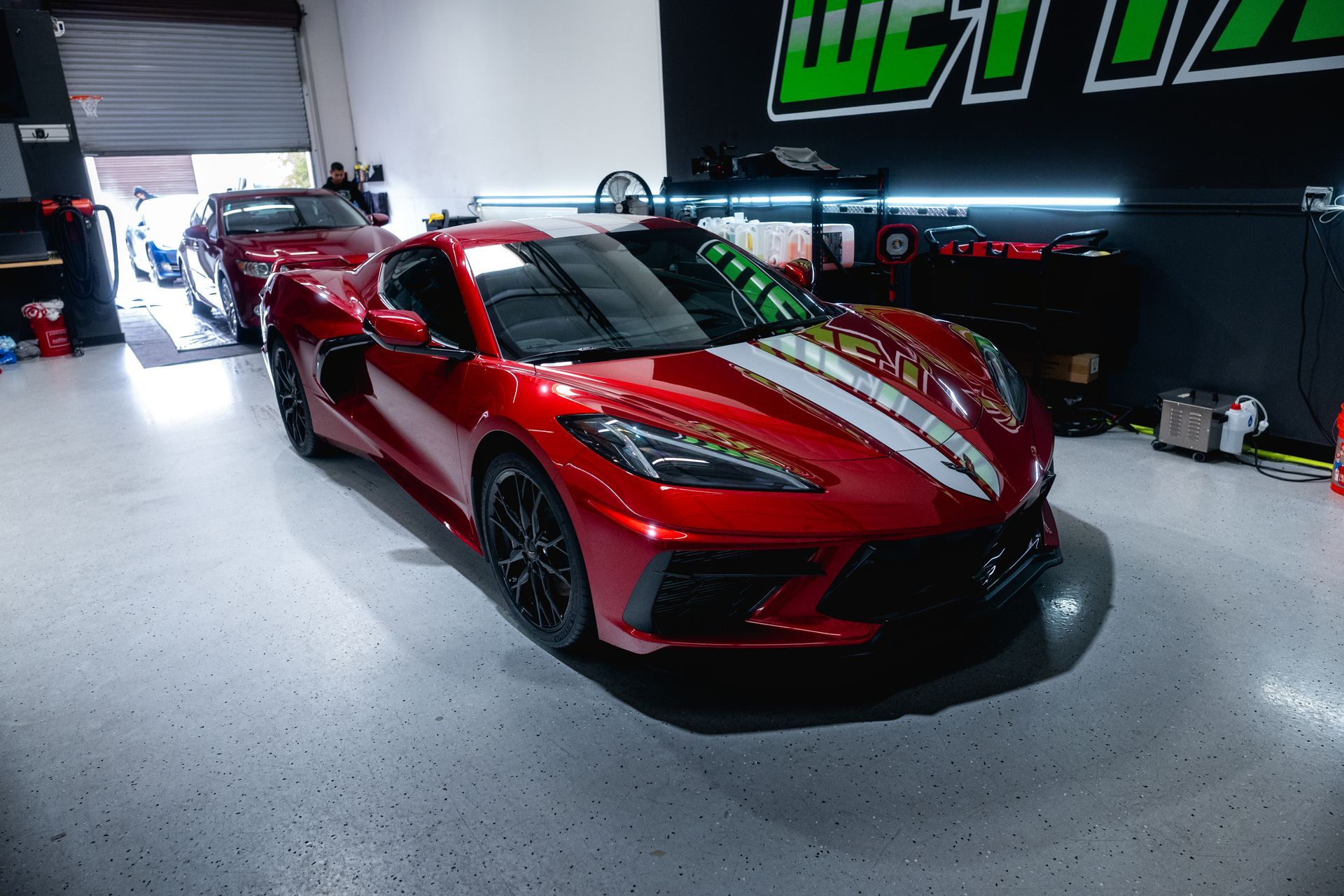 Red Corvette sports car with white stripes in a garage. Another car is partially visible in the background.