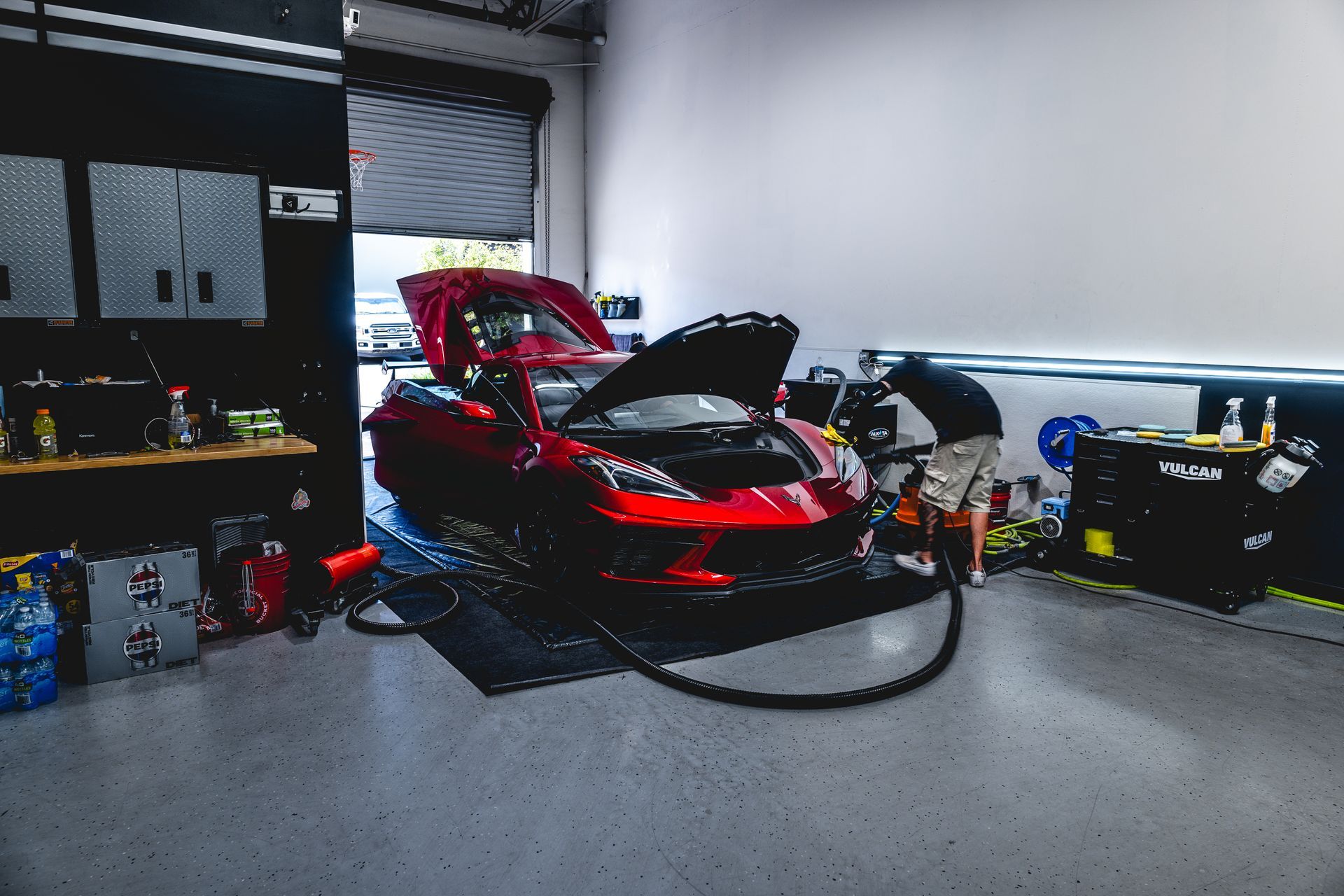 Red sports car in a garage with hood open, being worked on. A person works near it.