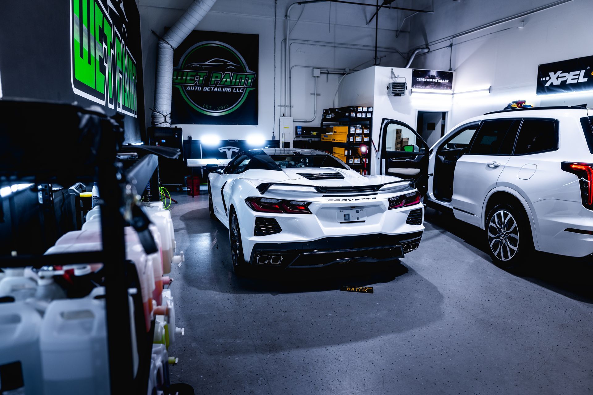 White Corvette and SUV in a detailing shop, lit by bright overhead lights.
