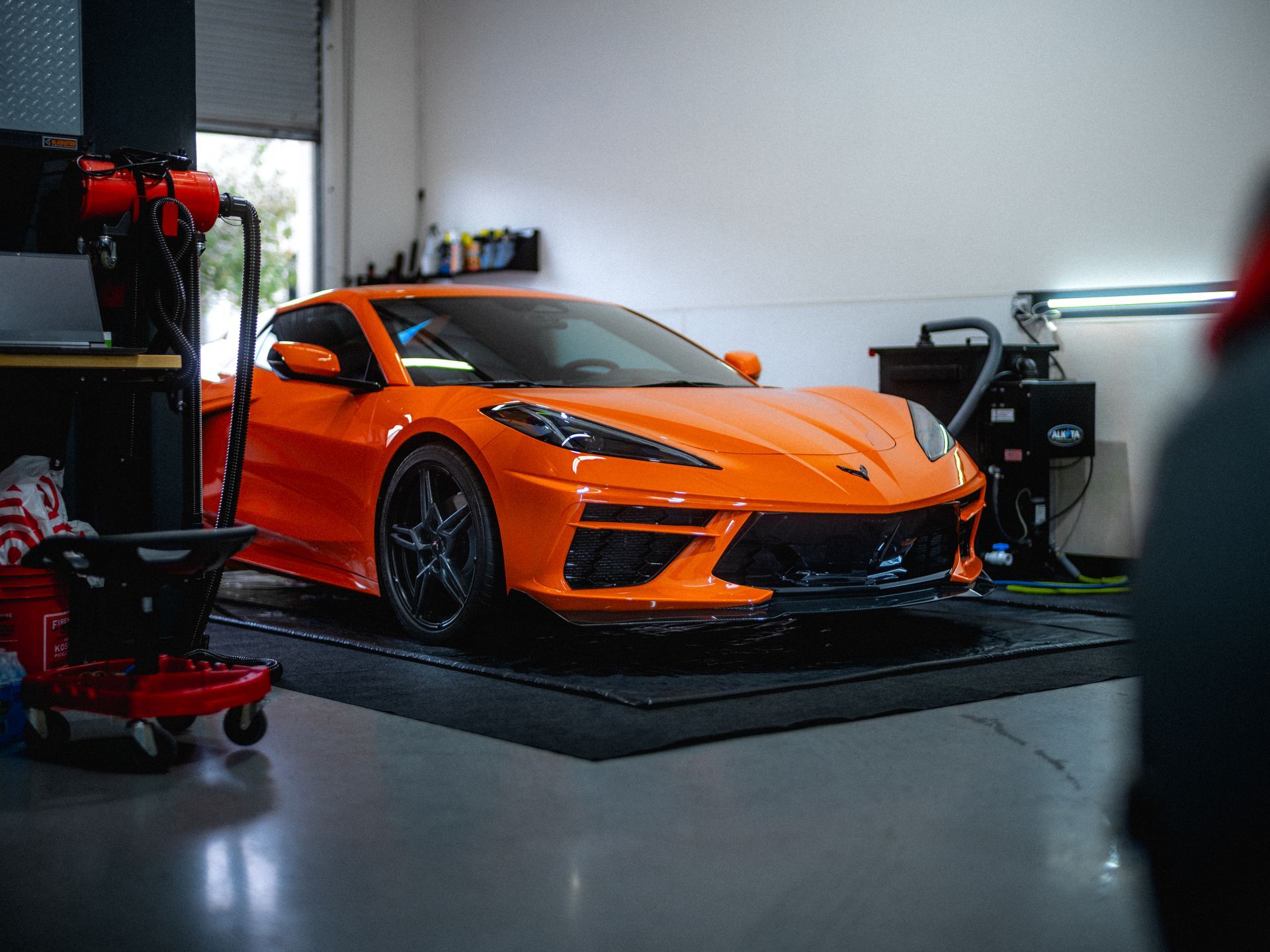 Orange Corvette sports car in a garage, on a black mat, with tools and a person visible.