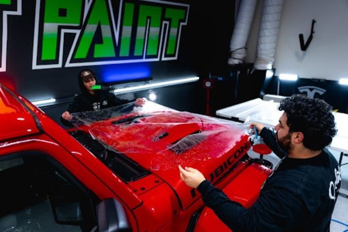 Wet Paint technician performing PPF installation on a red Jeep Wrangler hood under bright shop lights 