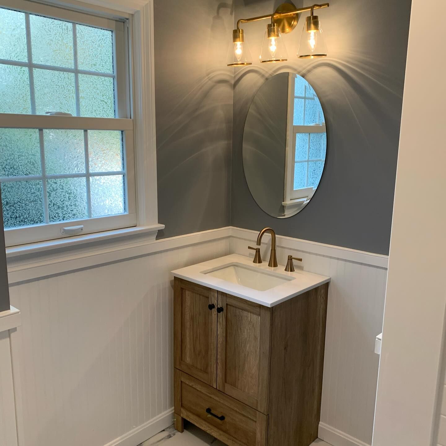 Bathroom with a wooden vanity, oval mirror, gold fixtures, and a window.