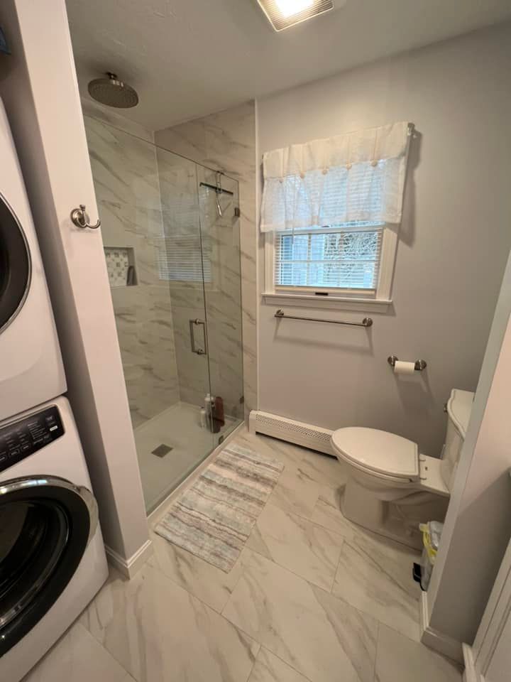 A modern bathroom with a glass shower, a toilet, and a stacked washer/dryer on the left. White marble-look tile.
