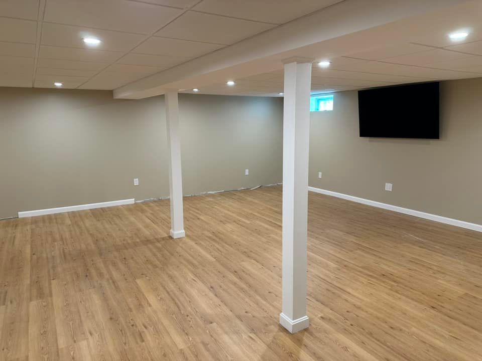 Finished basement with wood flooring, tan walls, white support columns, and a mounted TV.
