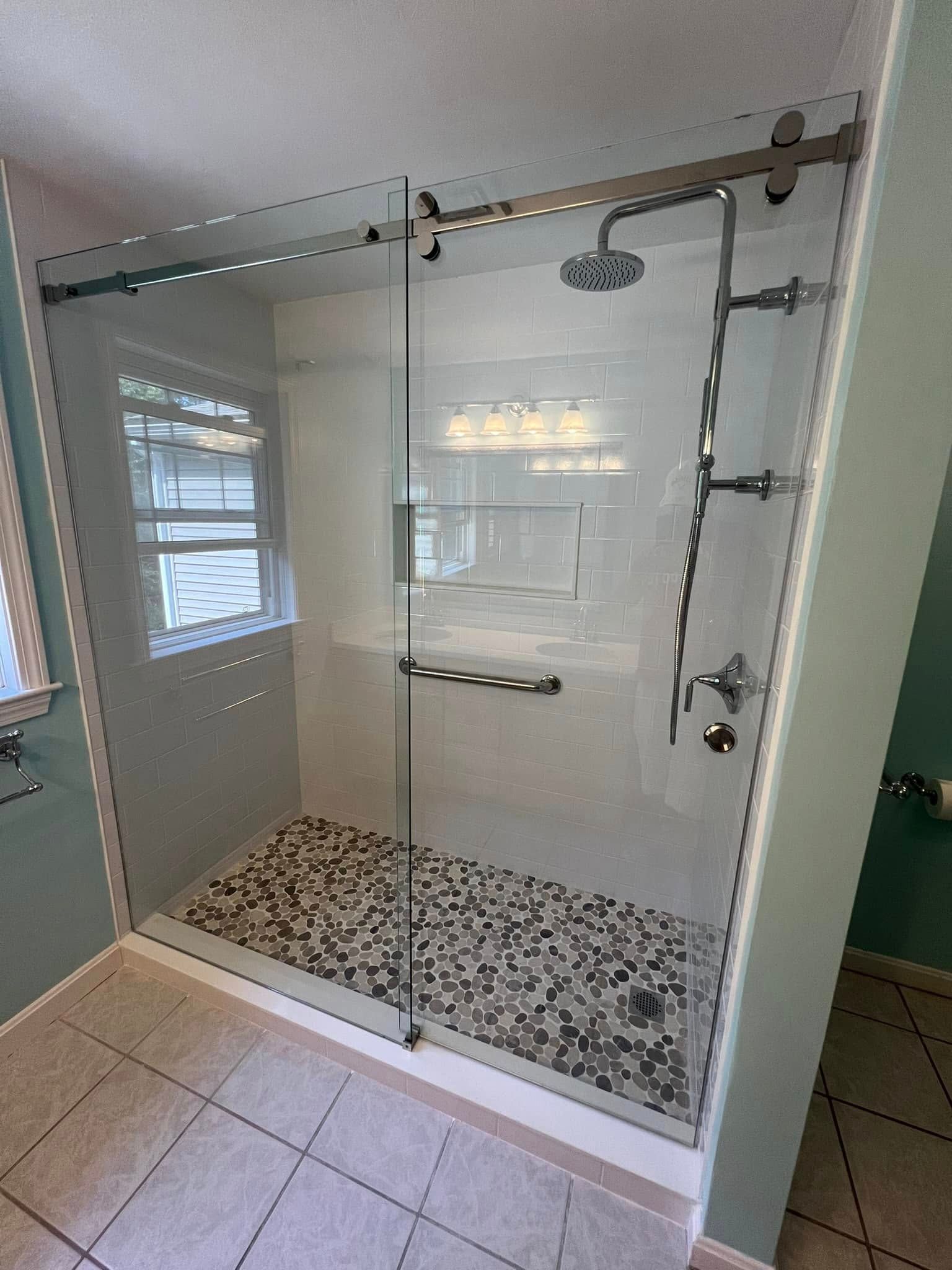 Modern glass shower with pebble floor, sliding door, chrome fixtures, white tile walls, and a window.