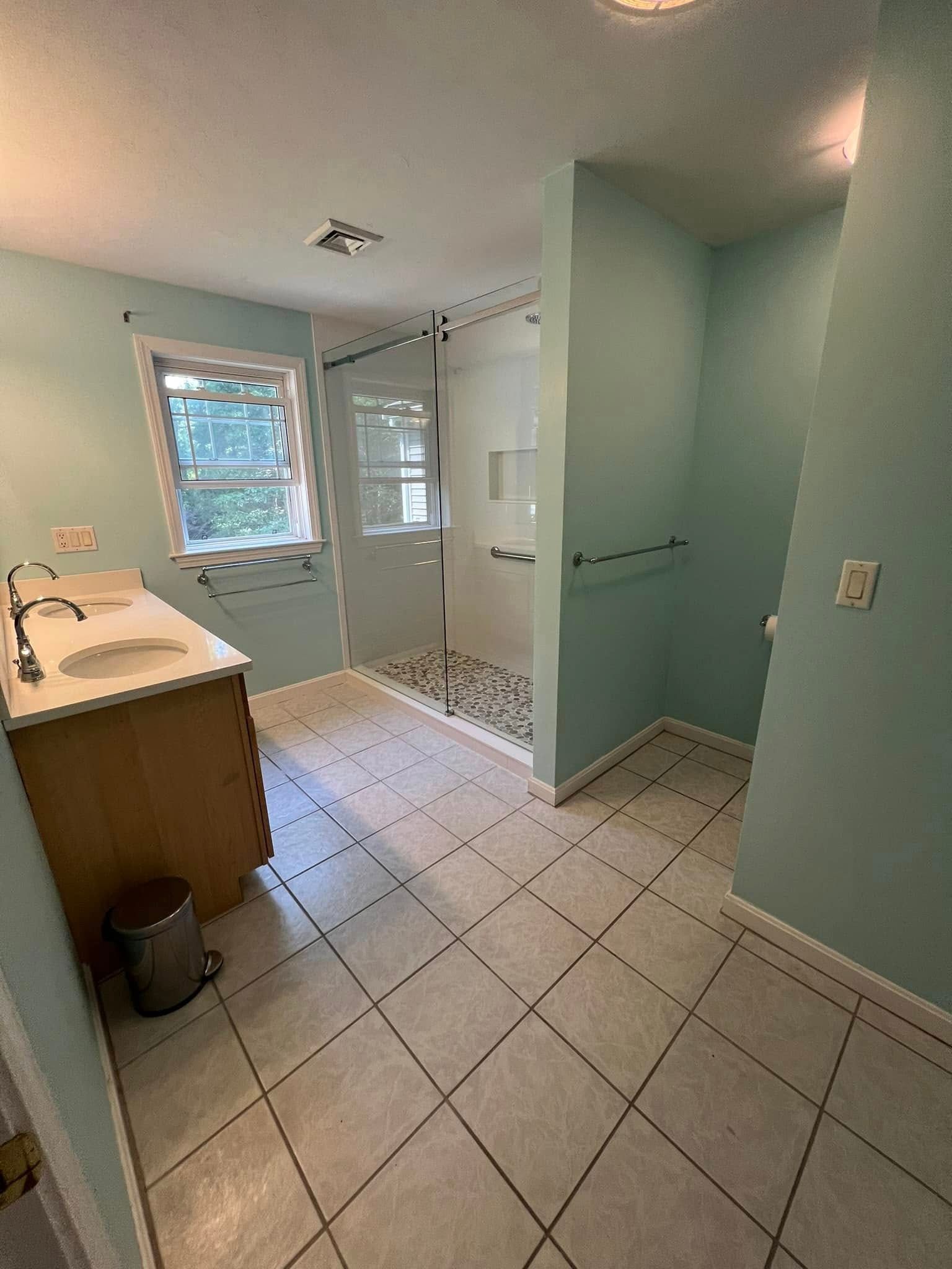 Bathroom with light blue walls, tan vanity, tiled floor, glass shower, and window.