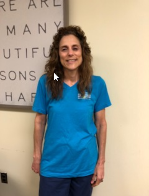 Woman with curly brown hair wearing a blue t-shirt and standing in front of a wall with words. She is smiling.