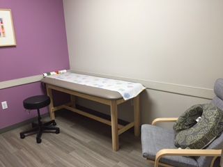 A medical examination room with a light-colored examination table, stool, and armchair. The walls are purple and beige.