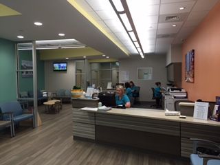 A brightly lit medical office reception area with staff working behind a desk. There are waiting chairs, colorful walls, and glass partitions.
