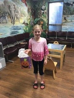 A young girl in a pink and purple shirt and black capri pants smiles, holding a Halloween bag in a waiting room with a jungle mural.