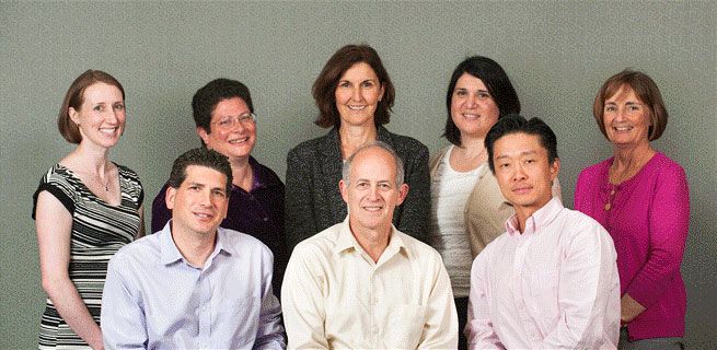 Group photo of nine people, mostly smiling, in front of a gray background. Diverse group, some in business attire.