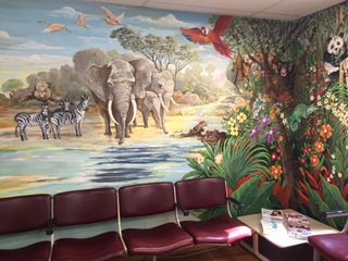 A waiting room with a mural of African animals and lush vegetation, including elephants, zebras, and a macaw, above burgundy chairs.
