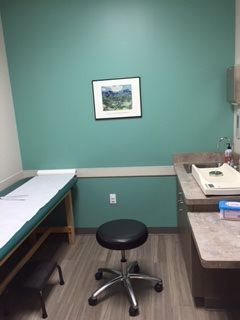 Examination room with an aqua wall, exam table, and stool. A framed picture hangs on the wall, with a sink area to the right.