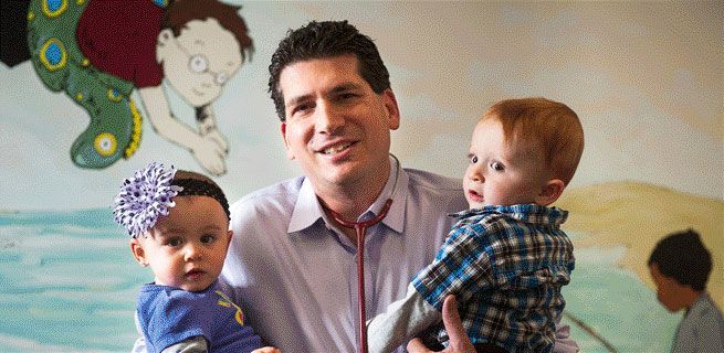 A doctor smiles while holding two babies. A mural is visible in the background.