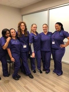 Six healthcare workers in blue scrubs pose for a group photo in an office setting. They are smiling.