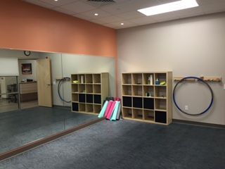Empty dance studio with large mirror, two cubby shelves, exercise hoops, and colorful rolled mats. Peach and beige walls, dark blue floor.