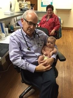 A doctor, holding a baby, looks at the camera in an office setting. A smiling woman is in the background.