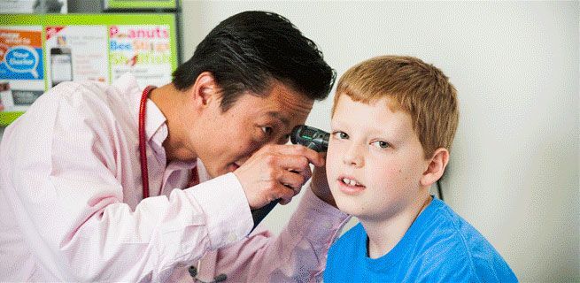A doctor uses an otoscope to examine a young boy's ear. The boy wears a blue shirt and looks concerned.