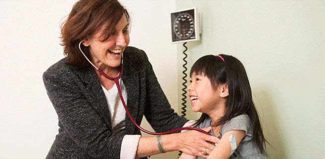 A doctor uses a stethoscope on a young girl in a medical setting. Both are smiling.