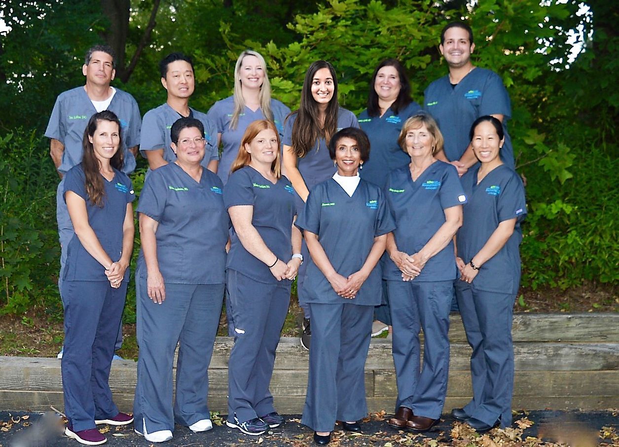 Group of diverse medical professionals in blue scrubs posing outside; smiling, trees in the background.