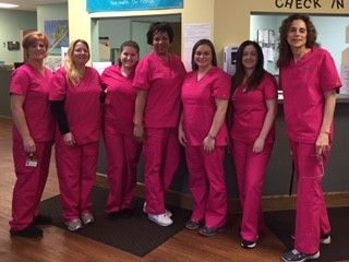 Seven healthcare workers in matching pink scrubs stand behind a check-in desk. They appear to be smiling in a clinical setting.