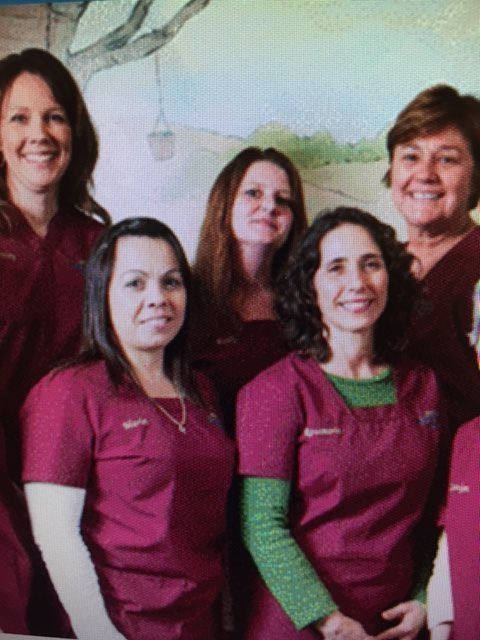 Group of six women in burgundy scrubs smiling, posing together indoors in front of a mural.