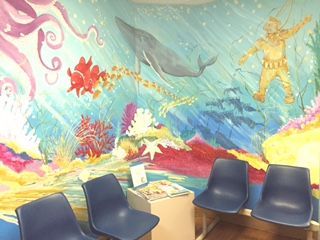 A waiting room with blue chairs and an underwater-themed mural featuring a whale, fish, and figure with raised arms.